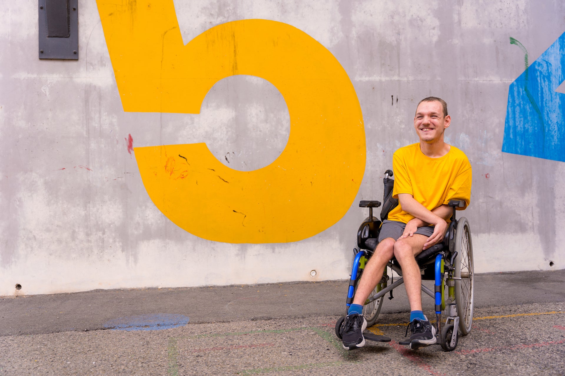 A person wearing a yellow shirt and sitting in a wheelchair, is in front of a concrete wall with yellow and blue paint that could be large numbers.